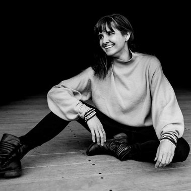 Boston elopement photographer Korri Leigh Crowley sits on the ground in the Boston Seaport wearing black pants and tan sweater and smiles 