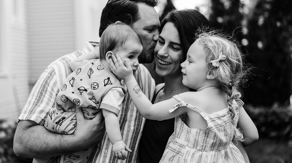 A family of four embraces and smiles while the young daughter caresses the cheek of her infant brother