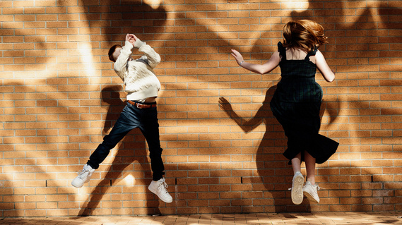 Two pre-teen children jump in front of a brick wall covered in bright orange reflections of light
