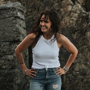 Korri Leigh Crowley in a white tank top and jeans smiles standing in front of large rocks on the beach