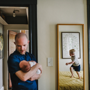 A father holds a newborn while his son jumps on the bed while reflected in a mirror
