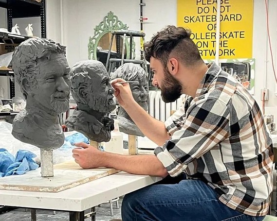 Douglas sculpting a clay bust on a table in-between two other clay busts.