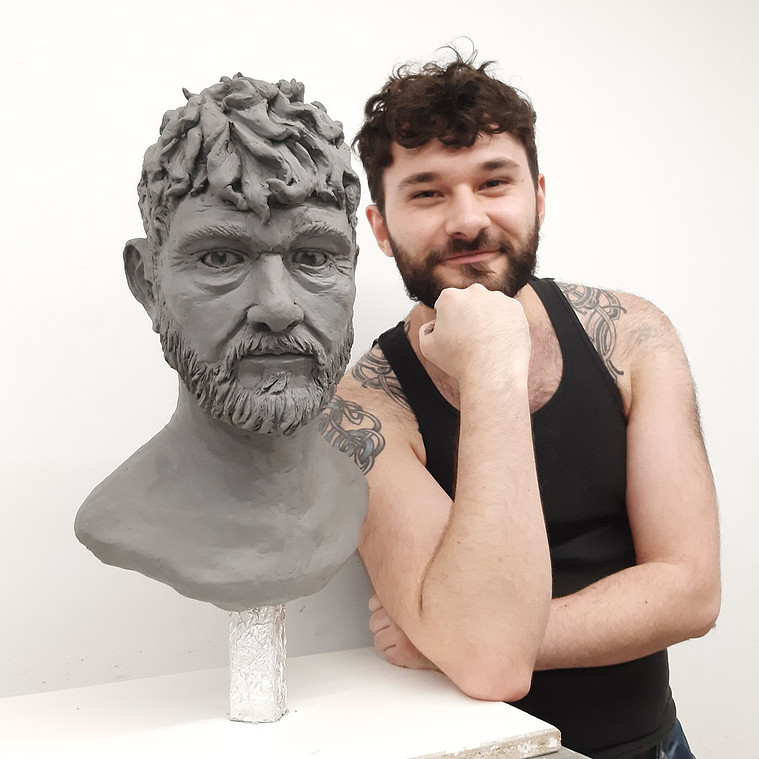 Douglas poses beside a clay bust of himself. He is smiling with one fist under his chin. 