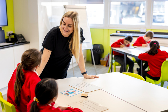 Female primary school teacher smiling while engaging with two young female students at their desk. In the background, three more students are focused on their classwork, illustrating an active and engaging classroom setting at St Peters at Gowts school
