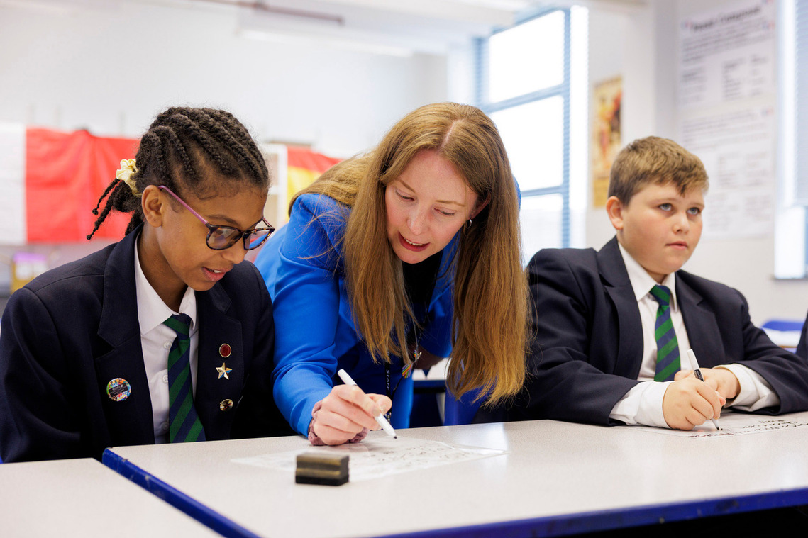 Female teacher at Park Vale Academy, Nottingham, leaning in with a pen to discuss a female student's work, while a male student looks towards the whiteboard, highlighting engaged learning.
