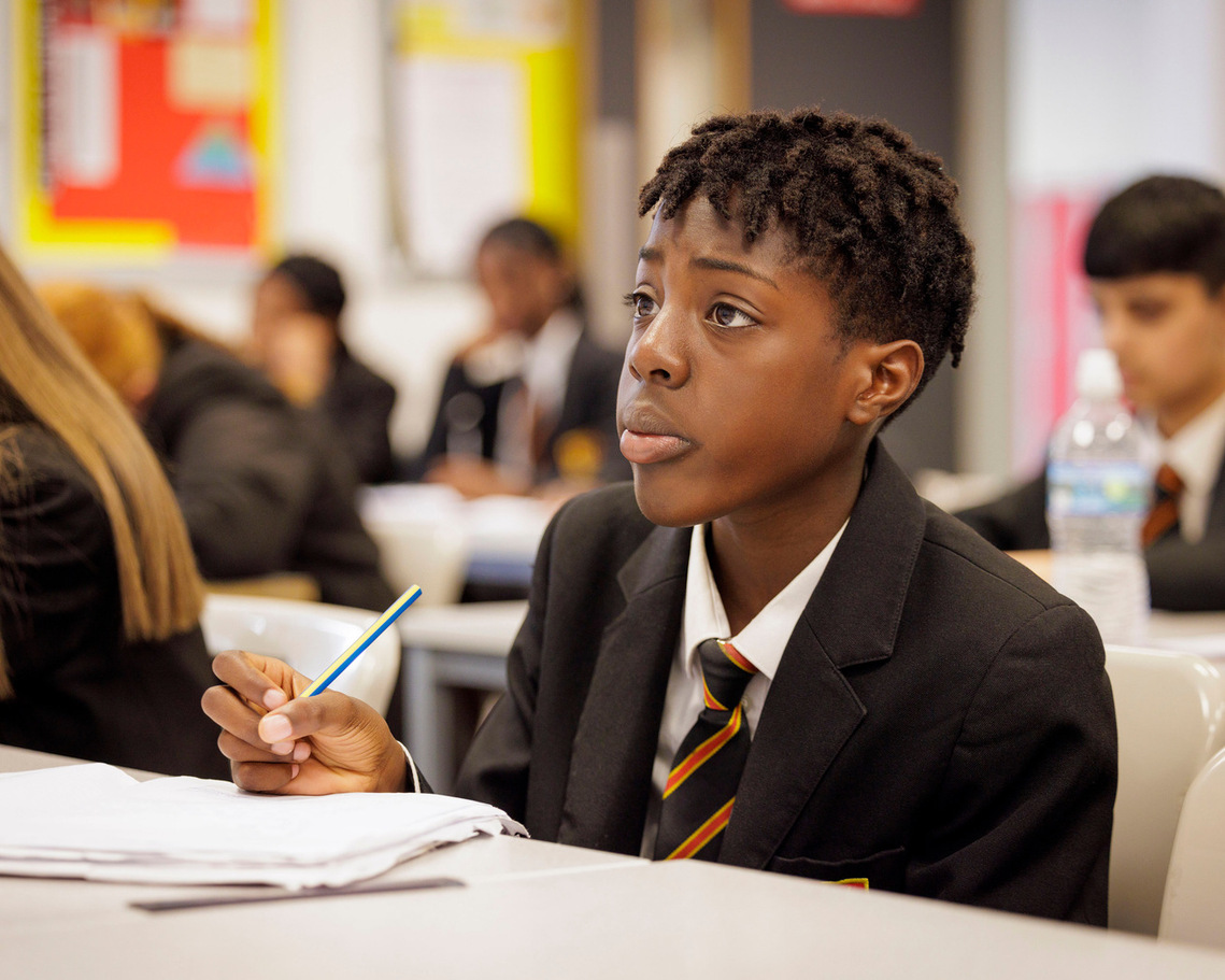 Senior school student at St Margaret Ward Catholic Academy, Stoke-on-Trent, focused on independent work. Male student is holding pencil, looking at the whiteboard, with a shallow depth of field highlighting concentration in the classroom.
