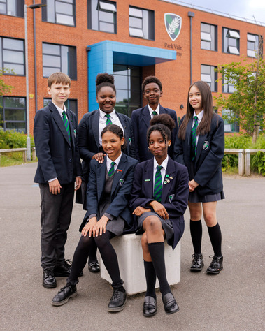 Six diverse secondary school students posing together, smiling proudly in front of Park Vale Academy's main site, representing a positive and inclusive school community.