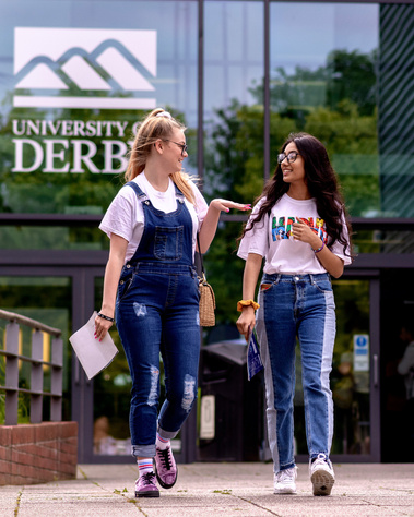Two prospective undergraduate female students walking together, engaged in conversation while holding a university brochure, exploring campus life and study options at the University of Derby.