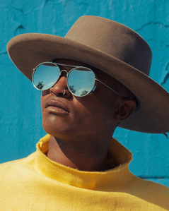 moody colorful portrait young black man wearing fedora hat and mirrored sunglasses and yellow top standing with blue background
