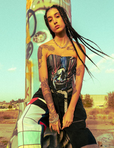 Young mixed race female model wearing corset with cartoon clown graphic on it checkered pants has tattoos long black hair stading in front of metal graffiti  covered pole and blue skies 