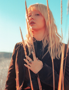 moody colorful portrait Blonde female model wearing black standing in high brush field with blue sky behind her
