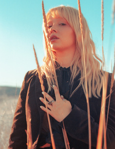 Blonde female model wearing black standing in high brush field with blue sky behind her