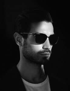 close up of male model face profile light skin short stubble beard brown hair slicked back wearing black sunglasses white t shirt sports coat blazer illuminated by high contrast light on part of face and shadowy background photograph william gormley