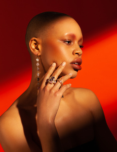 colorful orange background with a close up headshot of topless bald light skin young woman model Fitz looking to her left with right hand raised to her face wearing jewelry rings earrings designed by German kabirski, photograph by William Gormley