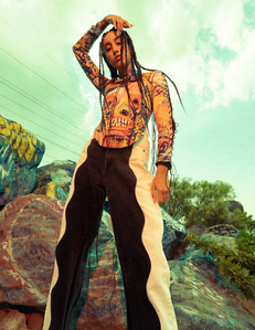 woman long hair weaves black white pants rusty corset art of Jean-Michel Basquiat arm raised above head reaching over left side while standing illuminated by colorful moody red light in front of large graffiti boulders and blue skies william gormley