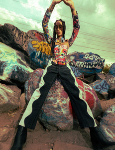 moody colorful portrait female model with dark hair wearing balck and white pants and corset with art print sitting on boulder rock covered in graffiti with legs spread wide and hand above her head
