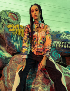 moody colorful portrait brunette female model sitting on graffiti covered boulder rocks with hands resting on lap wearing black and white pants and corset with sleeves illumiinated by red light and green skies