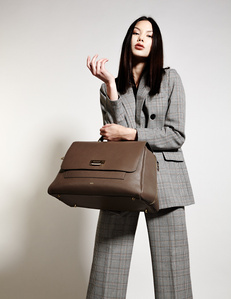 brunette female model long hair down dressed in gray business suit holding brown leather travel bag standing infront of plain white background photographed by William Gormley