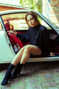 brunette caucasian female model wearing short black dress and black boots sitting inside white sports car with door open facing outward parked outside on brick driveway created by william gormley