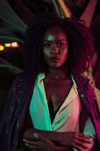 moody colorful portrait of black female model with arms crossed and black leather jack over shoulders and white button up shirt with her face and afro hair illuminated by pink and green light and palm leaves in background