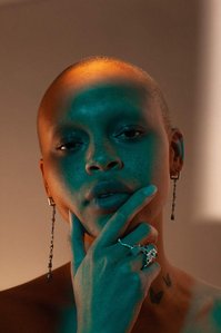 shadowy background illuminated blue face close up facing forward topless bald light skin young woman model Fitz right hand raised to chin wearing jewelry big rings long earings designed by german kabirski, photograph by william gormley