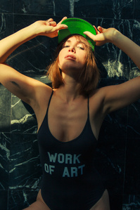 moody colorful portrait of Female model dressed in black bathing suit that says work of art with arms raised above head touching bill of hat standing against black marble title wall with ambient light covering the partial top half of her body