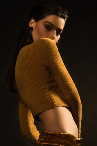 moody colorful portrait female model named ruby king wearing a gold long sleeve top with standing with her right shoulder facing forward partially blocking some of her face standing in front of dark background, photograph by william gormley