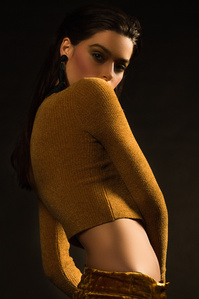 female model named ruby king wearing a gold long sleeve top with standing with her right shoulder facing forward partially blocking some of her face standing in front of dark background, photograph by william gormley
