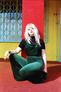young female model with blonde hair wearing green overalls crouching squatting in front red pillar