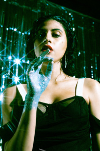 moody colorful portrait medium close up of Vartuhi woman model brown hair light skin styled with black dress white shoulder straps standing facing forward raising right hand covered in silver glitter in front of sparkly silver streamer filled backdrop