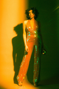 Vartuhi female model brown hair light skin wearing colorful orange green sequin jump suit sandal heels standing facing foreward in front of warmly lit shadow moody filled half backdrop, photograph William Gormley