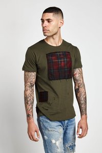 male model with short brunette crew cut hairstyle with arms covered in tattoos wearing an olive green t shirt that has a square plaid pattern patch stitched on front and wearing blue denim jeans with a white background for e commerce catalog