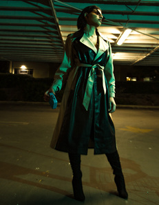brunette female model standing in underground parking garage lot wearing long black and tan leather trench coat with black boots illuminated by green and yellow ambient light.