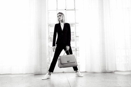 blonde female model wearing black suit and white sneakers holding leather taupe hand bag standing in front of white curtains and window photograph by william gormley