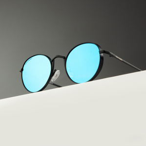 Round black rimmed mirrored blue lens sunglasses sitting on a white surface with a gray background in soft light