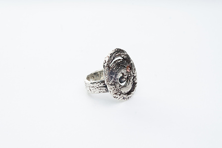 Textured silver ring in three-quarter view on neutral white ground.  The face of the ring features a flattened spiral with a textured surface.  A dusty pink cubic zirconia is set flush with the spiral surface, right of centre.
