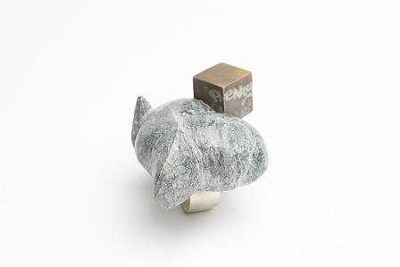 A ring with a thick, brushed silver band is pictured at 3/4 view on a neutral white ground. The top of the ring is set with an oversized rock carved in an abstracted mass.  At one edge, there is a small silver cube, patinated with the word "plenty".