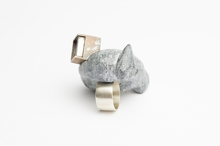 A ring with a thick, brushed silver band is pictured at an angle on a neutral white ground. The top of the ring is set with an oversized rock carved in an abstracted mass.  At one edge, there is a small silver cube, patinated with the word "plenty".