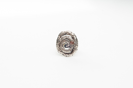 Textured silver ring in top view on neutral white ground.  The face of the ring features a flattened spiral with a textured surface.  A dusty pink cubic zirconia is set flush with the spiral surface, right of centre.
