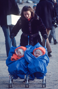 The young mother walking in the centre of Paris pushes twins in a double stroller; both children are wearing red hats.