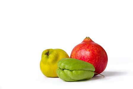 This is a still-life composition featuring a vibrant green chayote, a deep red pomegranate, and a bright yellow quince arranged artistically against a neutral background.