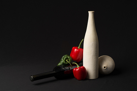 Still life featuring two white earthenware bottles and a black glass bottle, surrounded by vibrant red peppers, set against a dark background.