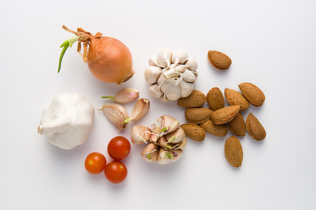 A still life composition featuring garlic, nuts, onions, and dwarf tomatoes arranged against a clean white background.