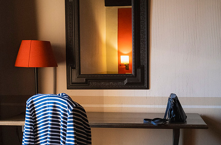 A quiet hotel room scene with a mirror, red lamp, and handbag on a small table—suggesting a moment of calm and stillness.