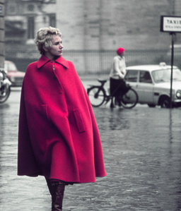 A fashionably dressed woman in a large red coat walks along a rain-soaked street in Paris while a man in a red cap holds a bicycle in the background.