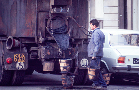 A worker at a tanker truck drains a bucket of asphalt to repair a hole in the road.