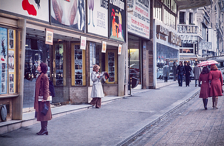 A street scene in Paris features the facade and entrance of a cinema. A woman stands observing the film listings while two more women in red coats share an umbrella nearby.