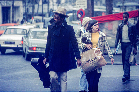 A street scene in Paris features a fashionably dressed man with a dark skin tone wearing sunglasses and a light-coloured Panama hat. He is carrying a violin case alongside a woman with a large plastic bag labelled &amp;amp;quot;Auburn&amp;amp;quot; crossing the street.