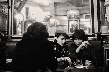 Two young women enjoy a lively conversation while smoking in a Montmartre café. A large mirror in the background reflects the warm atmosphere.