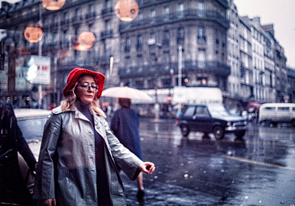 A busy intersection in Paris on a rainy day, with a woman in the foreground wearing a red hat and walking through the rain.